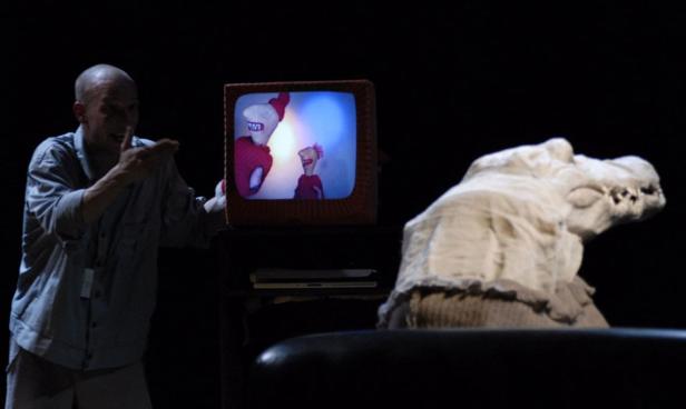 Ein Mann zeigt auf einen Fernseher mit animierten Figuren, neben ihm eine Krokodilskulptur.