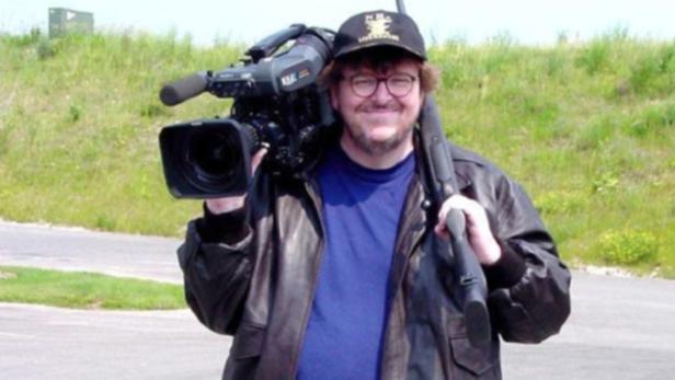 Michael Moore mit einer Kamera auf der Schulter und einem Gewehr im Arm.