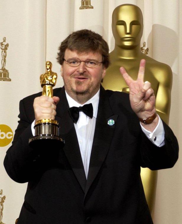 Michael Moore hält einen Oscar in der Hand und zeigt das Victory-Zeichen.