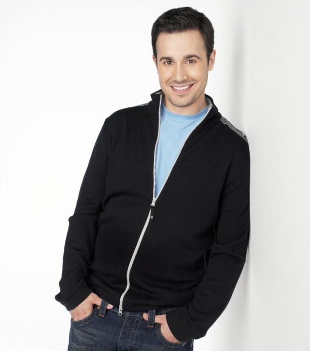 Ein lächelnder Mann in Jeans und Strickjacke lehnt an einer weißen Wand.