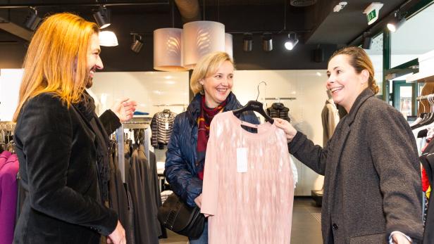 Drei Frauen in einem Bekleidungsgeschäft, eine hält ein rosafarbenes Oberteil auf einem Bügel.