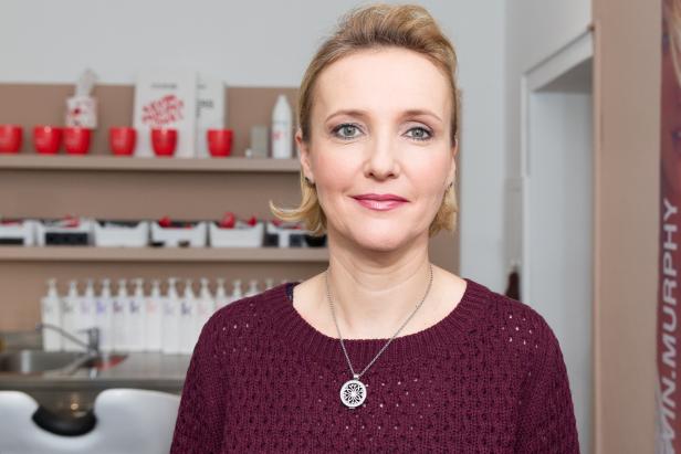 Eine Frau mit blonden Haaren und einem burgunderroten Pullover lächelt in die Kamera.