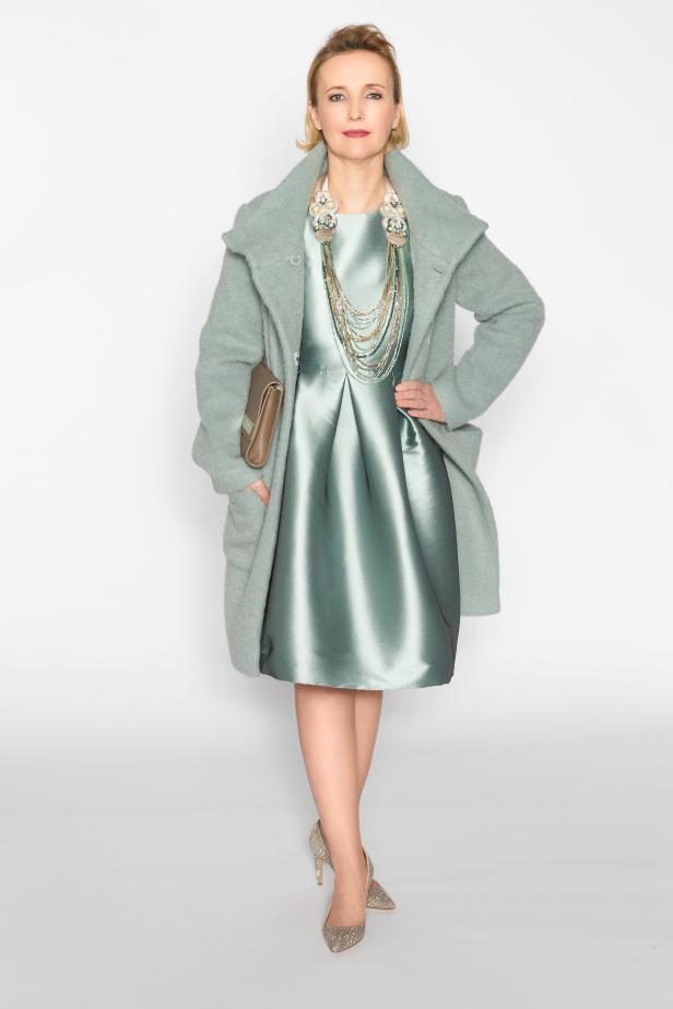 Eine Frau in einem grünen Kleid und Mantel posiert mit einer Hand in der Hüfte.