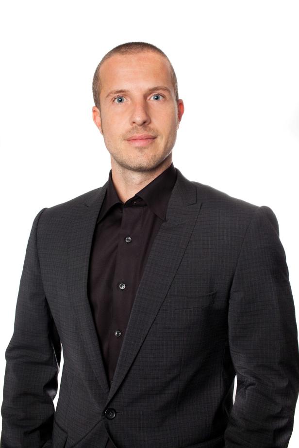 Ein Mann mit hellen Augen trägt einen dunklen Anzug und ein schwarzes Hemd.