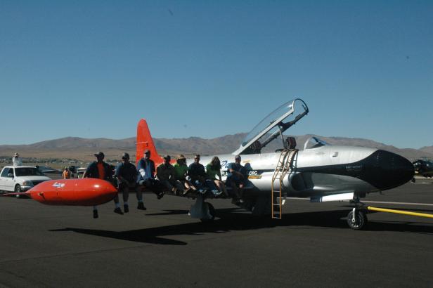 Eine Gruppe von Menschen sitzt auf dem Flügel eines silbernen Flugzeugs auf einem Flugplatz.
