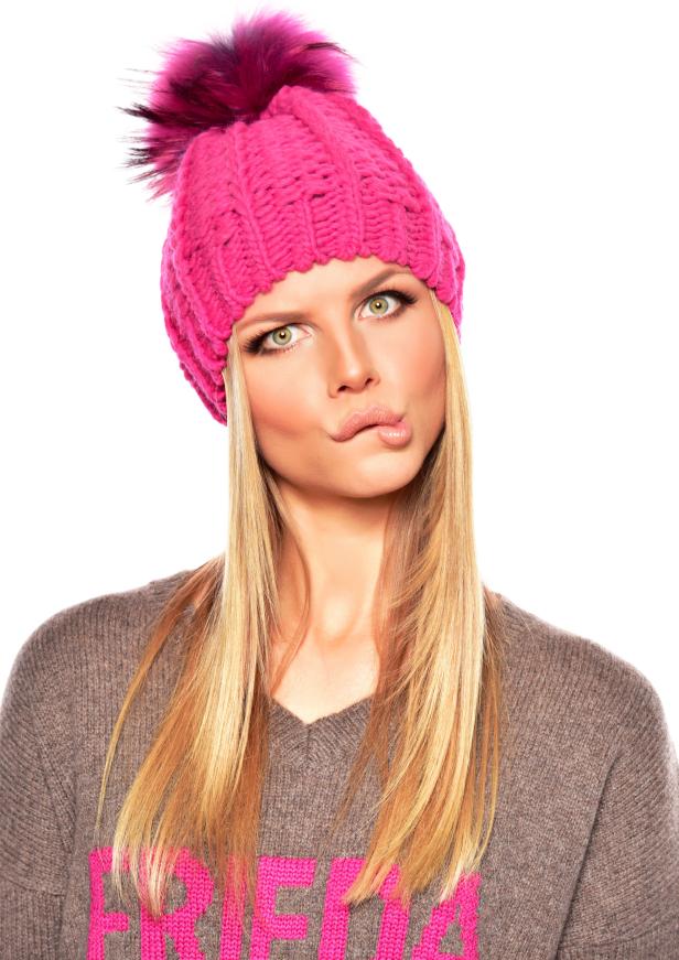 Eine blonde Frau mit einem pinkfarbenen Hut und einem grauen Pullover zieht ein Gesicht.