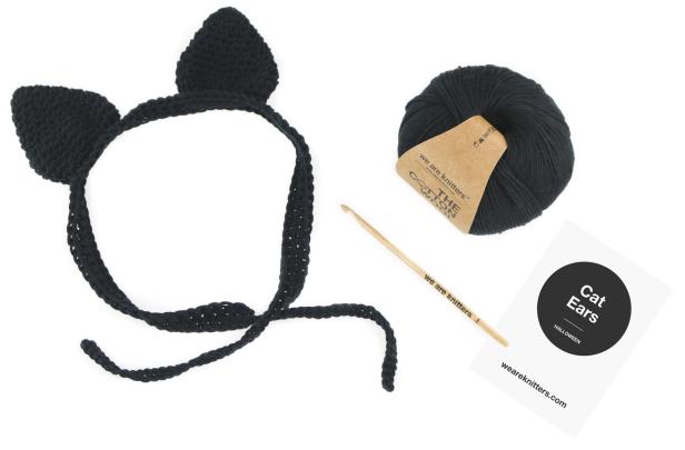 Ein Stirnband mit gehäkelten Katzenohren, eine Häkelnadel und ein Wollknäuel von We Are Knitters.