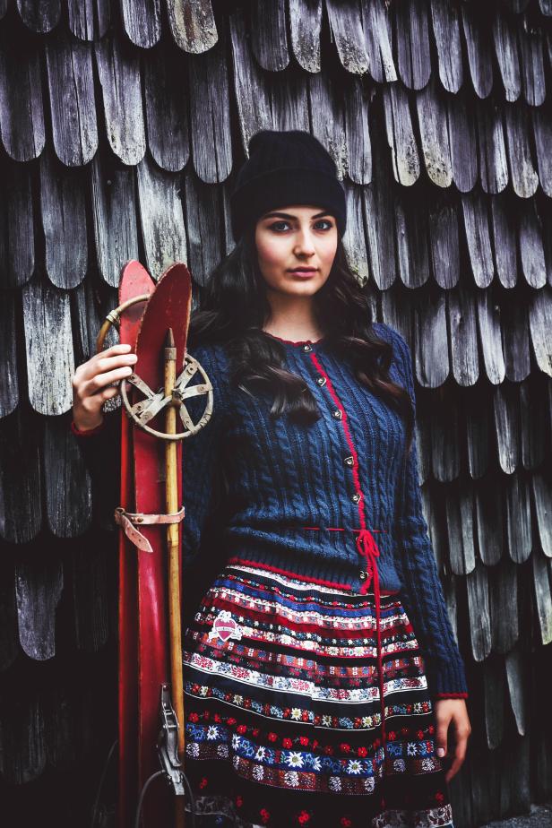 Eine junge Frau mit Mütze posiert mit alten Skiern vor einer Holzwand.