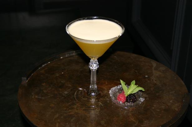 Ein gelber Cocktail in einem Martiniglas steht auf einem Tisch, garniert mit Beeren und Minze.