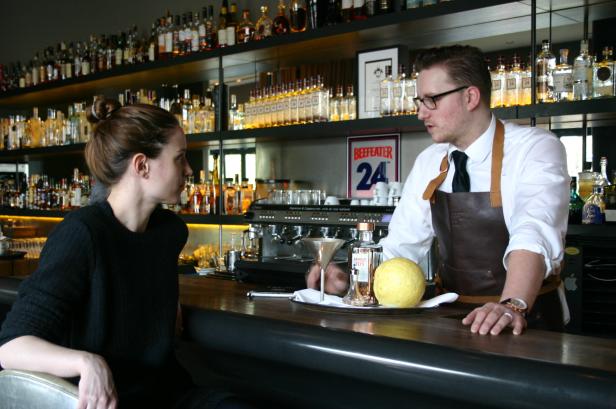 Eine Frau sitzt an einer Bar, während ein Barkeeper einen Drink zubereitet.