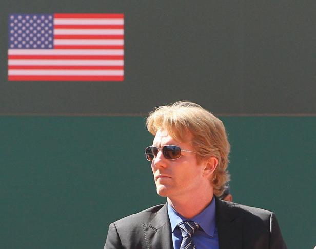 Ein Mann mit Sonnenbrille und blonden Haaren vor einer amerikanischen Flagge.