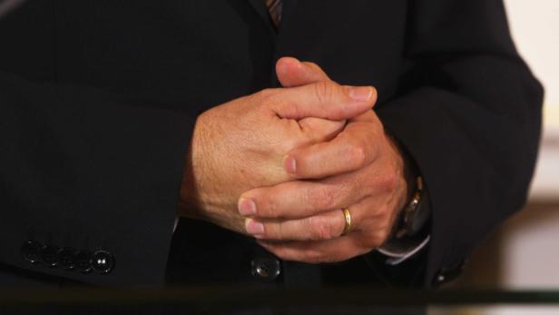 Ein Mann im Anzug hat die Hände gefaltet; er trägt einen Ehering.