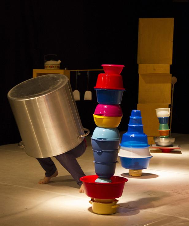 Eine Person balanciert einen großen Topf, während im Vordergrund bunte Schüsseln gestapelt sind.
