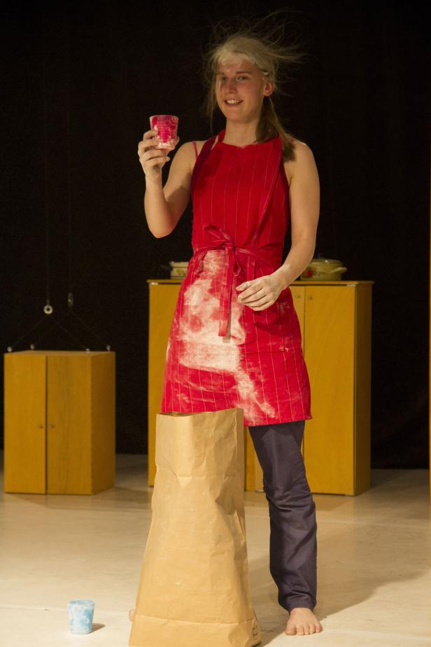 Eine Frau mit bemehltem Haar und roter Schürze hält ein Glas in der Hand.