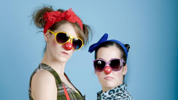 Zwei Frauen mit Clownsnasen und Sonnenbrillen posieren vor einem blauen Hintergrund.
