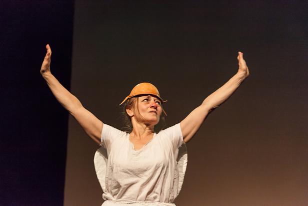 Eine Frau mit einem seltsamen Hut steht mit erhobenen Armen auf einer Bühne.