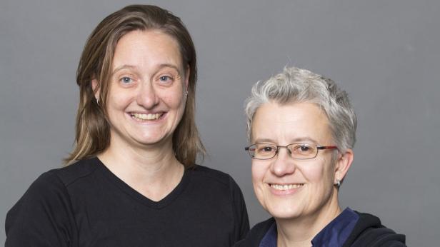 Zwei Frauen lächeln vor einem grauen Hintergrund in die Kamera.