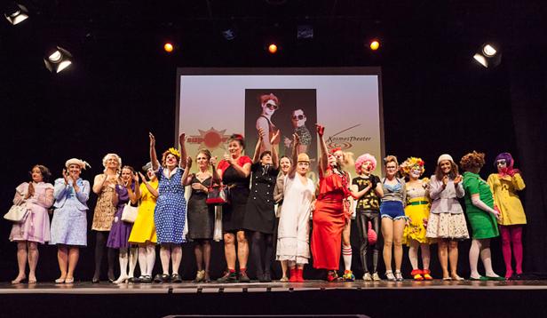 Eine Gruppe von Frauen in Kostümen steht auf einer Bühne vor einer Leinwand.