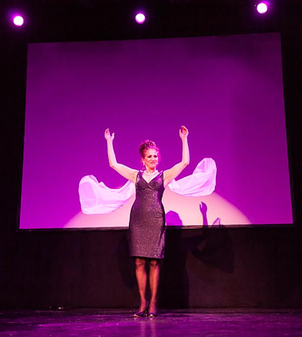 Eine Frau in einem Kleid steht mit erhobenen Armen auf einer Bühne vor einem lila Hintergrund.
