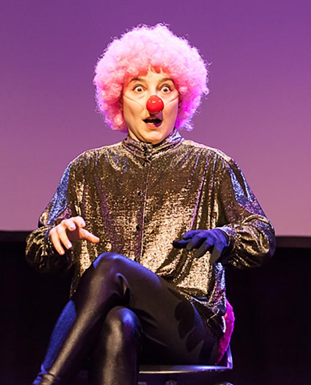 Eine Person mit rosa Perücke und roter Nase sitzt mit überraschtem Gesichtsausdruck auf einem Stuhl.