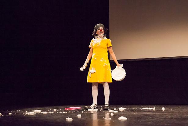 Eine Frau im gelben Kleid steht mit Sahne beschmiert auf einer Bühne.