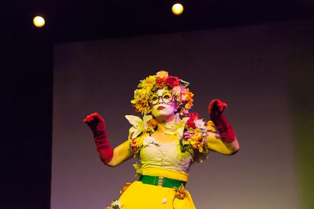 Eine Performerin mit Blumenkopfschmuck und roten Handschuhen steht auf einer Bühne.