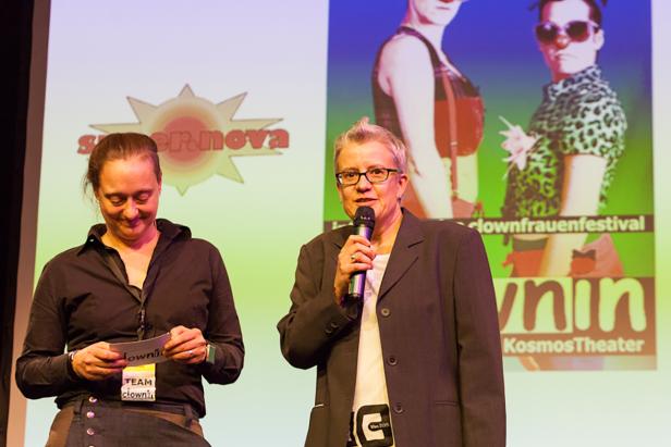 Zwei Frauen stehen auf einer Bühne vor einem Bildschirm des Clownfrauenfestivals.