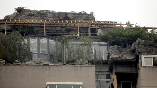 Ein Mann arbeitet an einer Holzkonstruktion auf dem Dach eines Hauses, das mit Felsen und Pflanzen geschmückt ist.