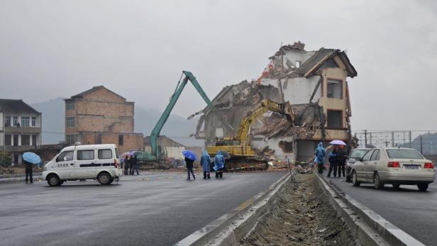 Ein Bagger reißt ein Gebäude in einer chinesischen Stadt ab, während Menschen zusehen.