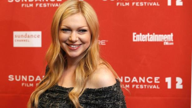 Eine Frau mit blonden, gewellten Haaren lächelt vor einem roten Hintergrund mit dem Logo des Sundance Film Festivals.