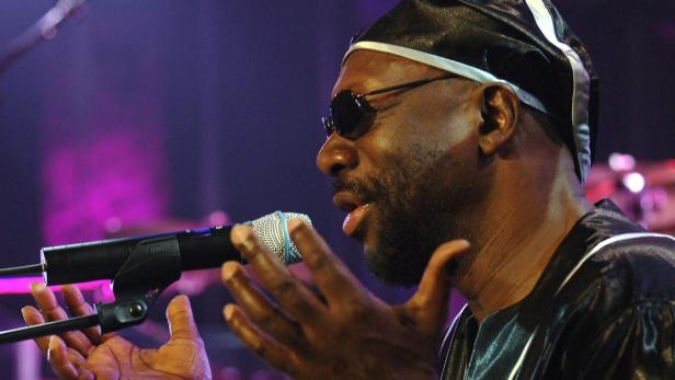 Ein Mann mit Sonnenbrille und Mütze singt in ein Mikrofon.