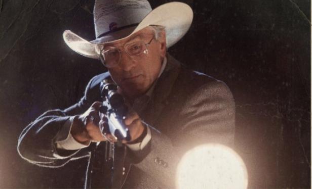 Ein Mann mit Cowboyhut und Brille zielt mit einem Gewehr.