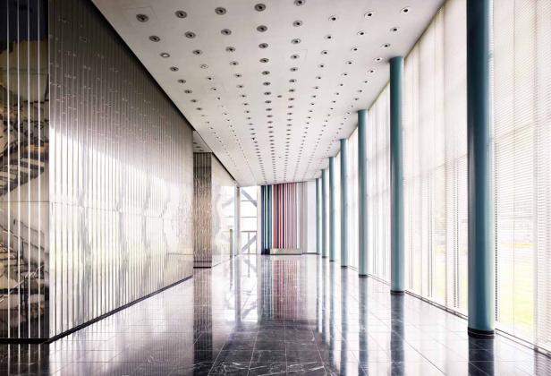 Ein langer Flur mit poliertem Marmorboden, Säulen und einer Wand mit vertikalen Metallstreifen.