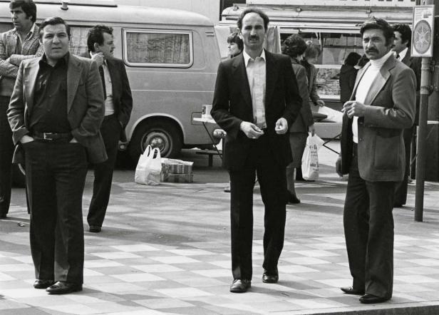 Drei Männer in Anzügen stehen auf einem Platz in Schwarzweiß.