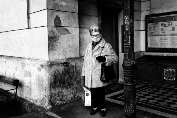 Eine ältere Dame mit Brille und Pelzmantel steht vor einem Gebäude.