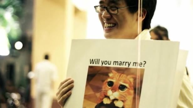 Ein lächelnder Mann hält ein Schild mit der Aufschrift „Will you marry me?“ und einem Bild von Kater aus dem Film „Der gestiefelte Kater“.