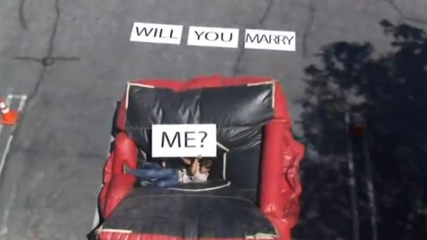 Eine Person liegt in einem roten Sessel und hält ein Schild mit der Aufschrift „Will You Marry Me?“.