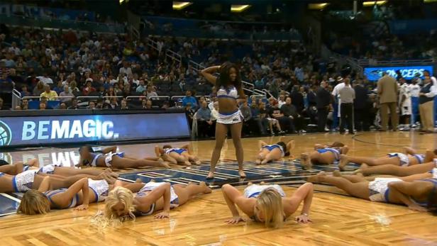 Eine Cheerleaderin steht vor einer Gruppe von Cheerleadern auf dem Boden in einer Arena.