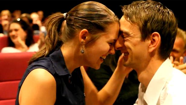 Ein verliebtes Paar berührt in einem Kinosaal die Stirn aneinander und lacht.