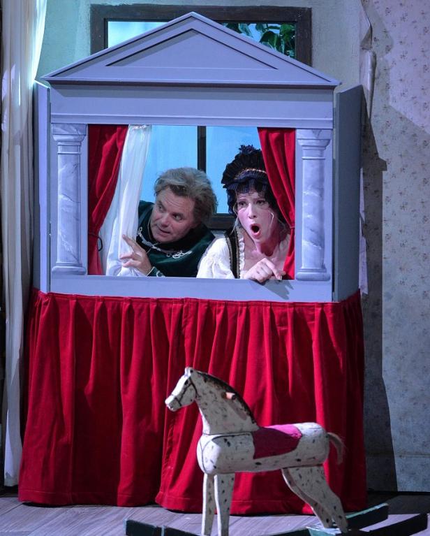 Zwei Schauspieler blicken überrascht aus einem Puppentheater, im Vordergrund ein Schaukelpferd.