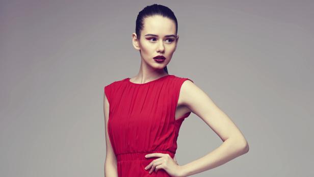 Eine Frau mit roten Lippen und rotem Kleid posiert vor einem grauen Hintergrund.