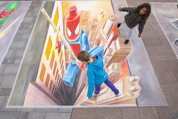 Zwei Kinder posieren auf einem 3D-Straßenkunstwerk mit einem Lego-Superhelden in einer Stadtlandschaft.