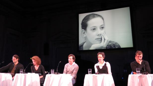 Fünf Personen sitzen an Tischen auf einer Bühne vor einem großen Bildschirm mit einem Porträt.