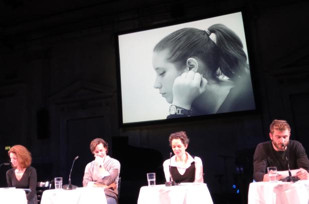 Vier Personen sitzen an Tischen vor einer Leinwand mit dem Porträt einer jungen Frau.