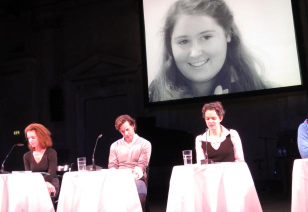 Vier Personen sitzen an Tischen auf einer Bühne, im Hintergrund ein Porträt auf einer Leinwand.