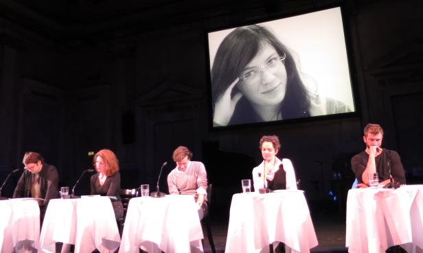 Fünf Personen sitzen an Tischen auf einer Bühne, im Hintergrund ein Porträt auf einer Leinwand.
