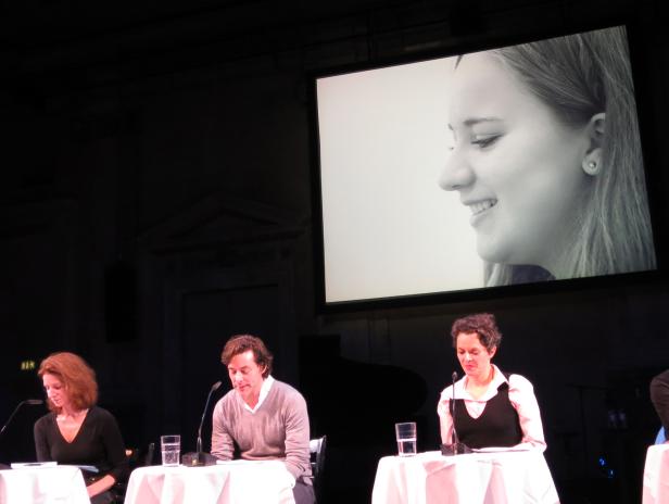 Drei Personen sitzen an Tischen vor einer Leinwand mit dem Porträt einer jungen Frau.