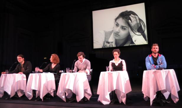 Fünf Personen sitzen an Tischen auf einer Bühne vor einer Leinwand mit einem Schwarzweißporträt.