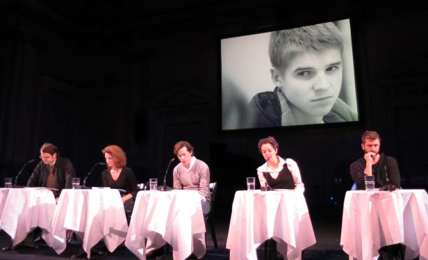 Fünf Personen sitzen an Tischen auf einer Bühne, im Hintergrund ein Schwarzweiß-Porträt eines jungen Mannes.