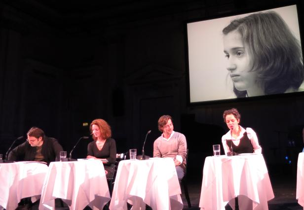 Vier Personen sitzen an Tischen auf einer Bühne, im Hintergrund ein Porträt auf einer Leinwand.